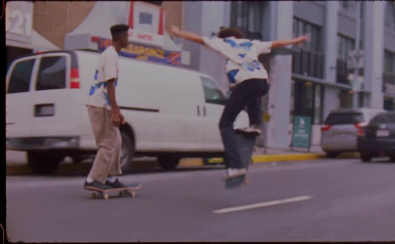 Two men with white t shirts and brown and black pants skateboard in the city street