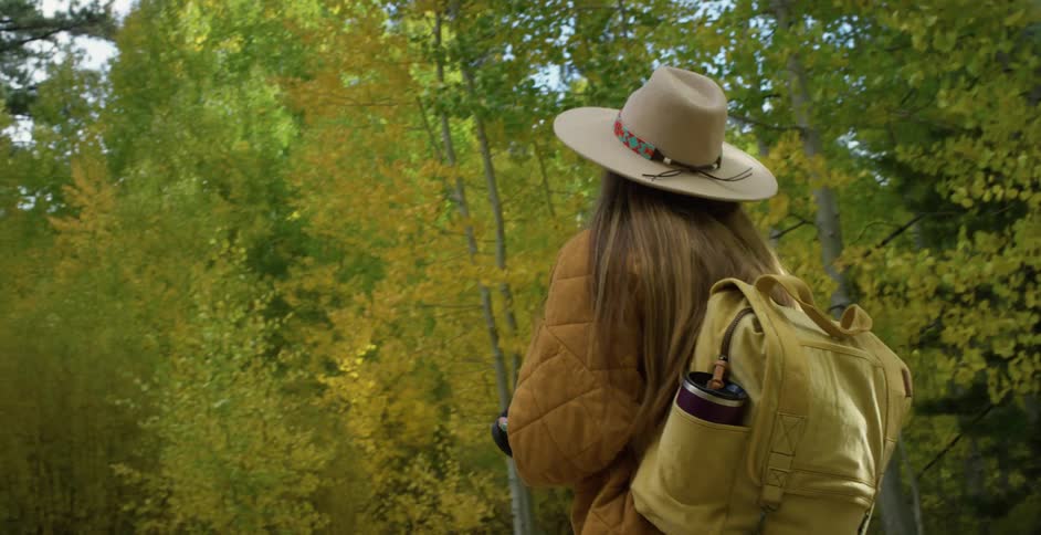 Woman with brown hat straight long hair coat and jeans carries camera andwalks down forest road in the woods