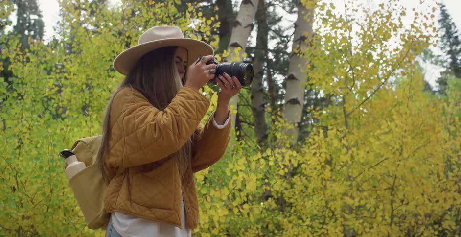 Woman with brown hat long straight hair jacket and backpack photographs trees in the woods
