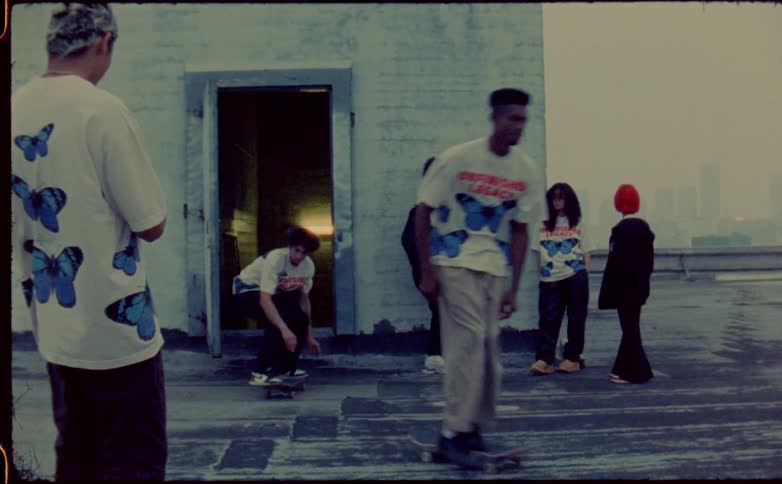 Friends stand and watch other friends skateboard out onto building roof as fog obscures city skyline