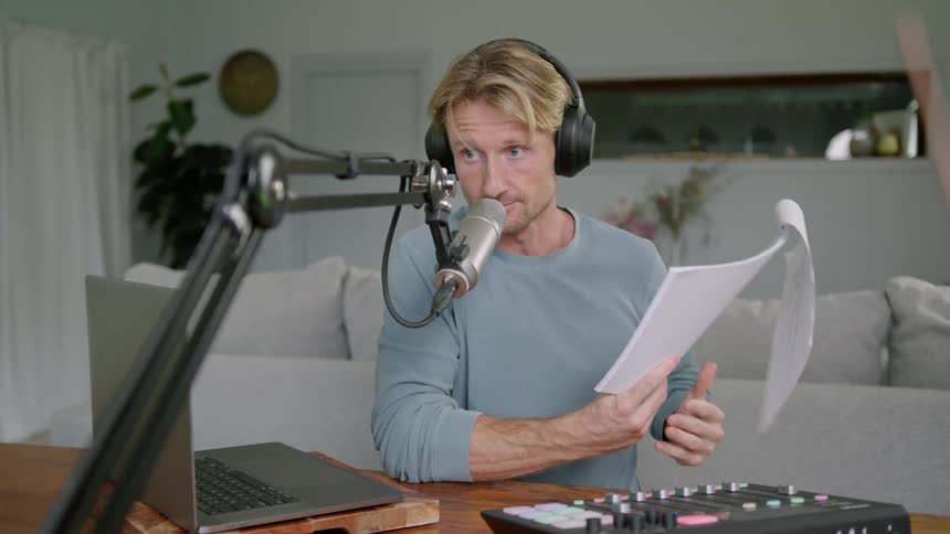 Man wearing headphones talks into a microphone near a laptop in a home studio