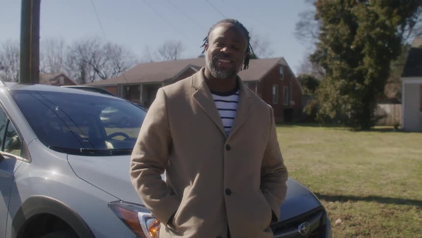 Black man wearing an overcoat smiling and standing in front of a car parked in front of a house at a residential neighborhood