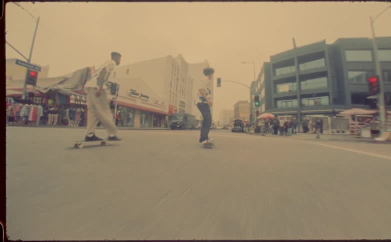 Vintage footage of two teenagers skateboarding through town in middle of road