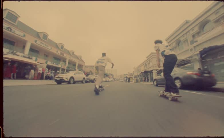 Two skateboarding males ride along middle of street between parked cars and stores