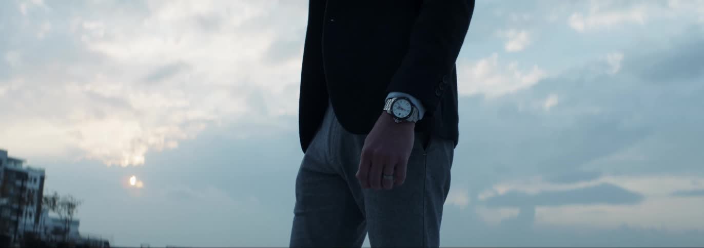 Man wearing blazer and slacks poses outdoors with apartment buildings in background as helicopter flies across sky