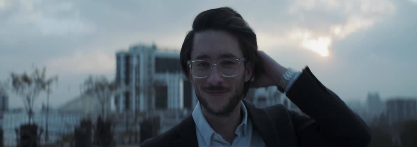 Man with glasses beard shirt and blazer jacket faces the camera smiling on rooftop with buildings in the background at dusk