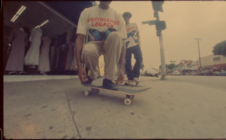 Two teenage boys skateboard outside of a clothing store on a street as cars drive past