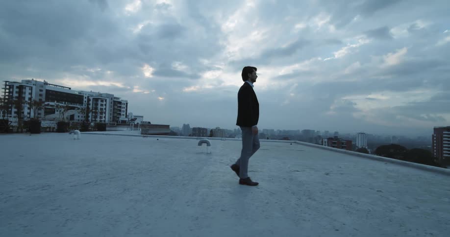 White man wearing formal clothing walking on rooftop of a building in a big city