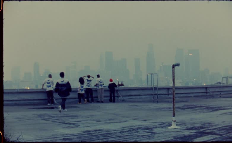 A group of teenagers and kids on a rooftop walk to the edge and look at the city skyline