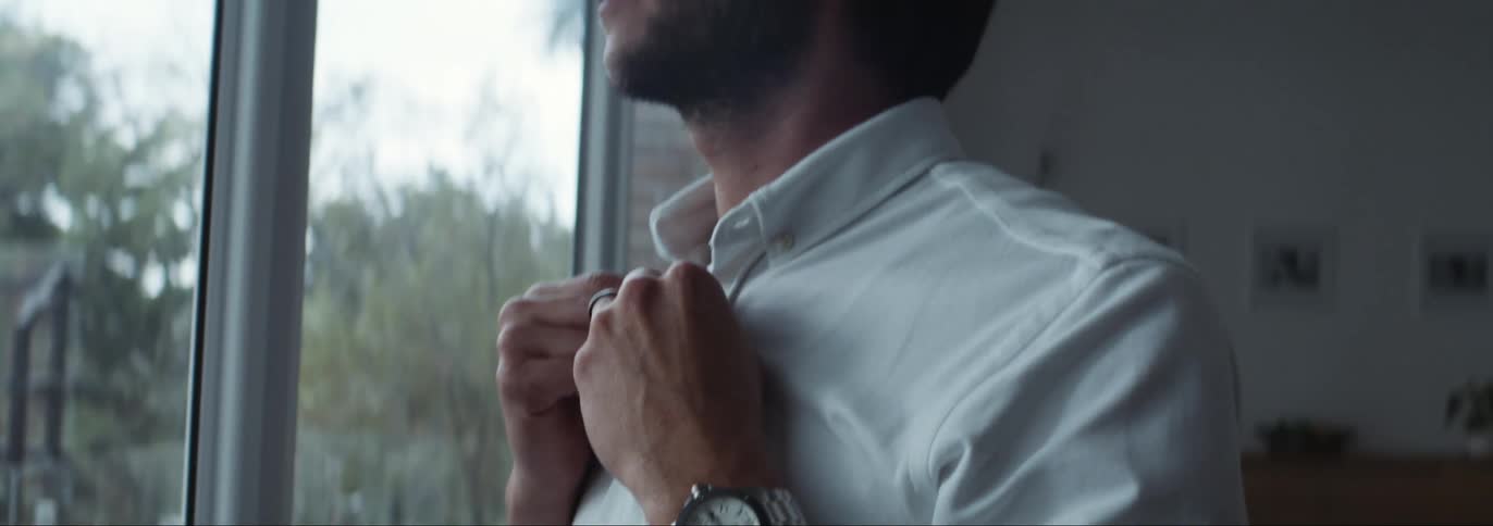 A man buttons his shirt by the window