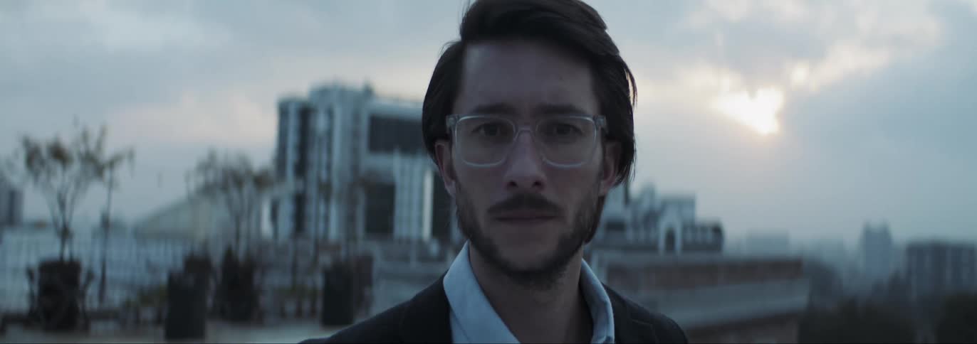 Man with glasses beard shirt and blazer jacket faces the camera smiling on rooftop with buildings in the background at dusk