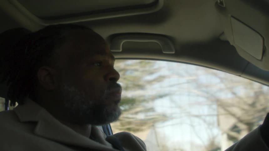 Man with dreadlocks beard and brown overcoat drives car on road in the neighborhood
