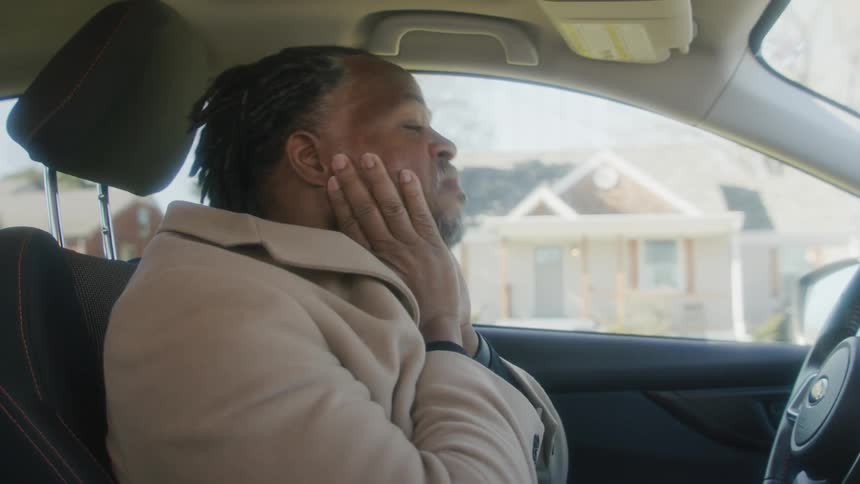 Nervous black man wearing an overcoat sitting at the drivers seat of his car parked in front of a house