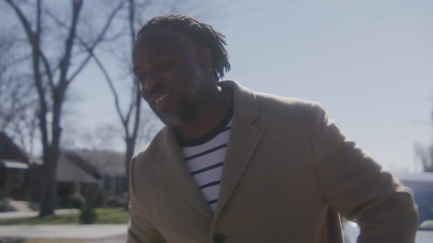 Black man wearing a jacket reaches out his hand and smiles then turns to walk past a parked car under a blue sky
