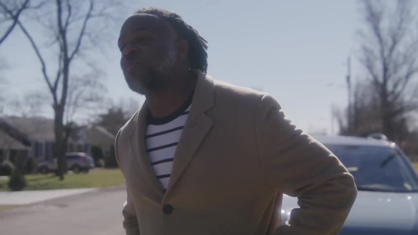 A man standing in a neighborhood street waves and speaks to someone