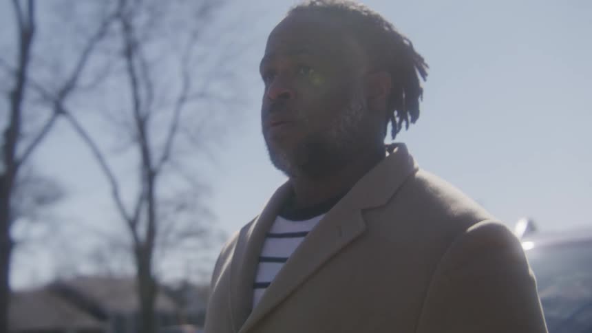 Man with dreadlocks beard and brown overcoat stands by car and looks around in the street
