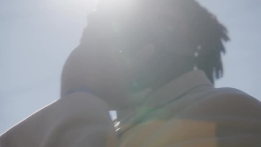 Man with dreadlocks beard and brown overcoat talks on smartphone under shining sun