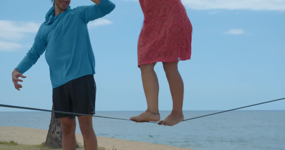 A man holds a womans hand as she walks across a tightrope on a beach
