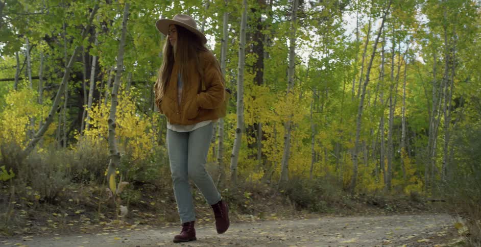 Woman with brown hat long straight hair jacket backpack and jeans walks on forest road in the woods