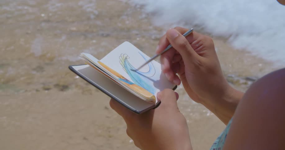 Woman wearing swimsuit on beach dips small brush in sea water to color wave in her small sketchpad with watercolors