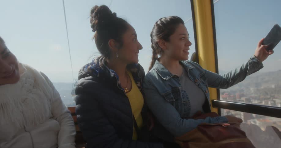 Three teenage girls ride in a gondola and one holds her cell phone and they pose for a photo