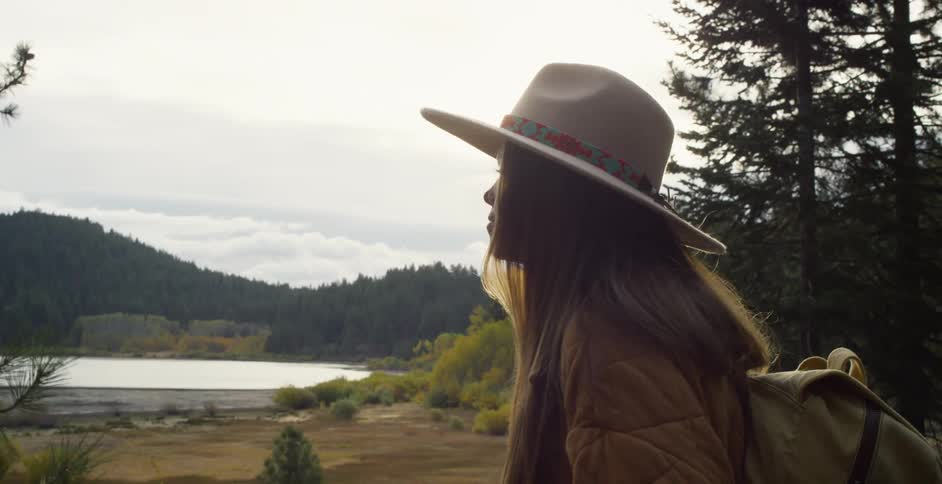 Woman with brown hat long hair jacket backpack and jeans walks on forest road in the woods