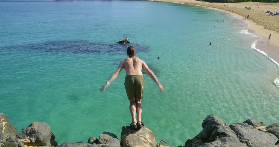 Man wearing green shorts standing on a cliff near a beach jumps on the water