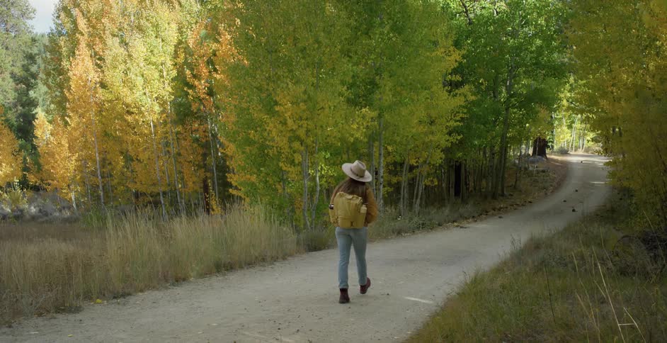 Woman wearing sun hat and carrying a backpack walks along narrow road through trees with fall foliage