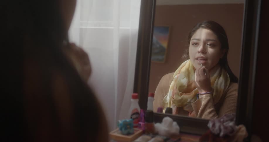 Woman with brown hair and beige sweatshirt and yellow scarf looks in mirror and puts on makeup