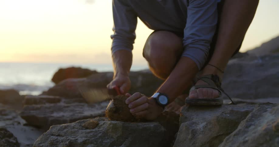 Man wearing sandals crouches on a rock near the ocean and tries to slice open a coconut with a knife