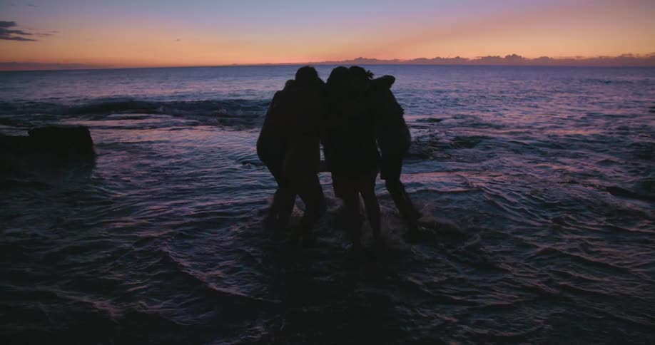 A group of men and women huddle together on the shore and turn in a circle as waves roll over their feet