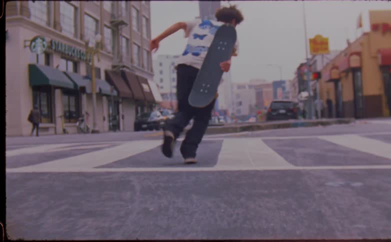 Skateboarder jumps of board and kicks it up to catch it as he keeps running across street
