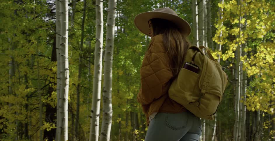 Woman with hat brown straight hair jacket backpack and jeans walks down forest road in the woods