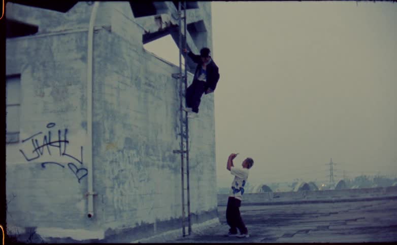 A man uses a smart phone to record his friend hanging from a ladder on a rooftop
