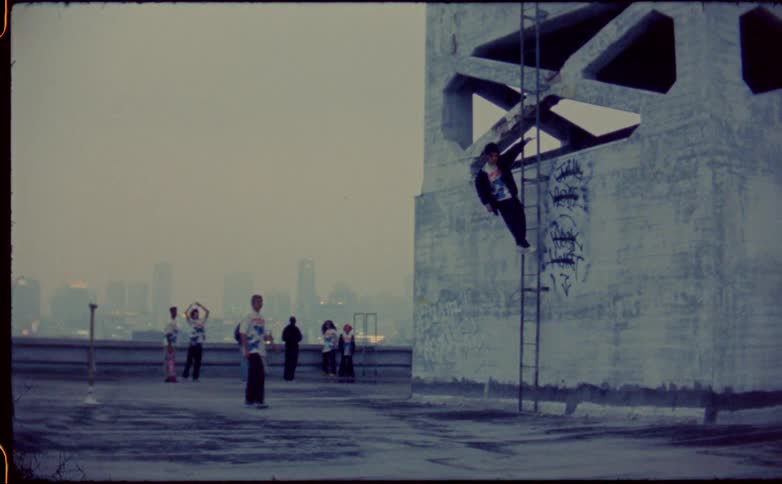 A group of teenagers hang out on a rooftop while one boy walks towards another boy who is hanging from a ladder against the wall