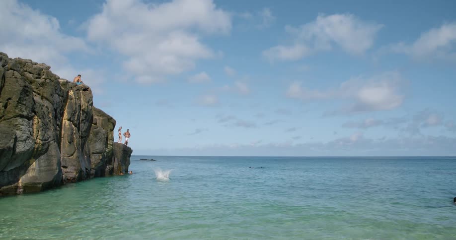 People standing at the edge of a cliff when one man jumps into the ocean on a sunny day
