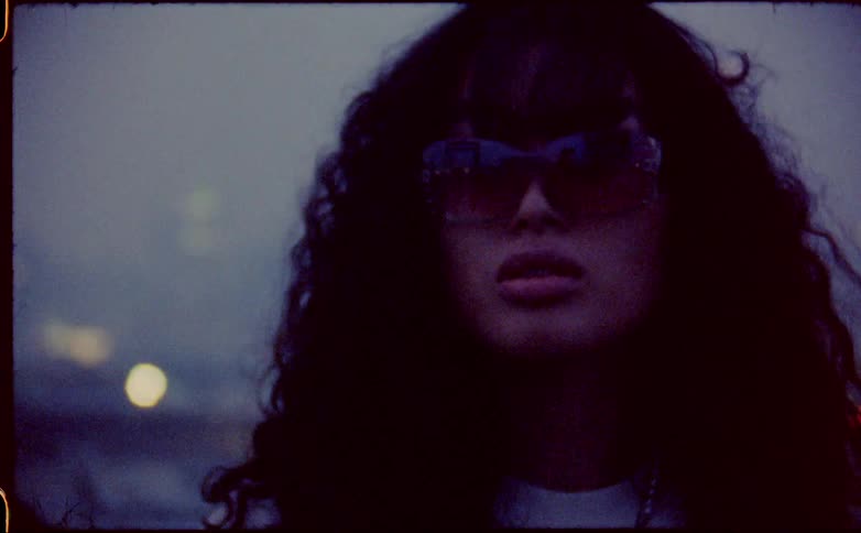 Young woman with curly hair wearing sunglasses near the city skyline covered in fog caught on vintage film