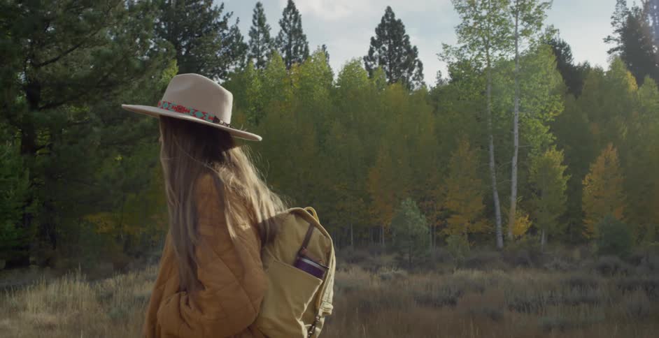 Woman with hat brown straight hair jacket backpack and jeans walks down forest road in the woods