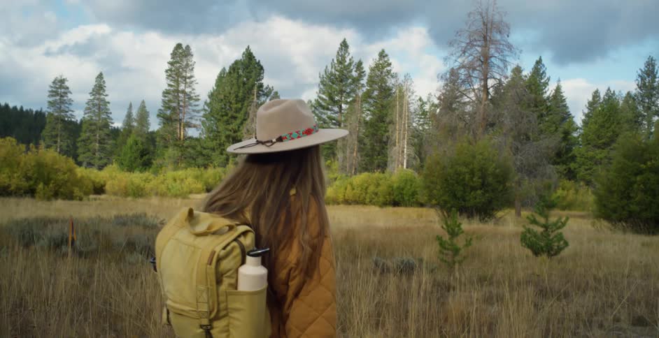 Woman with brown hat long hair jacket backpack and jeans walks around in the grassy field next to forest