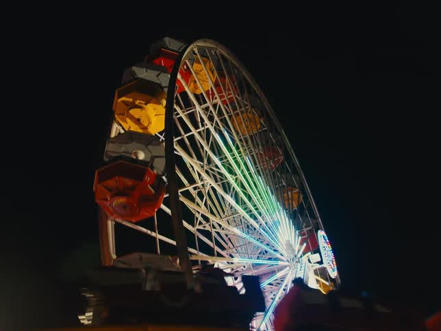 Ferris wheel with yellow and red seats hanging at its edge turns around in amusement park at night