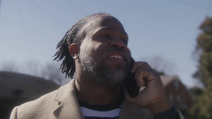 Black man stands outside in a neighborhood and holds a cell phone up to his ear