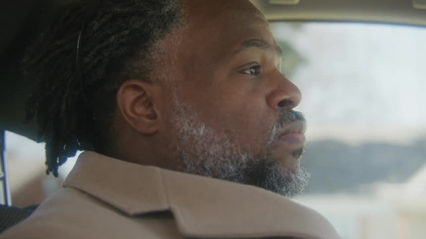 Man with dreadlocks beard and brown overcoat shakes his head in disappointment sitting in car