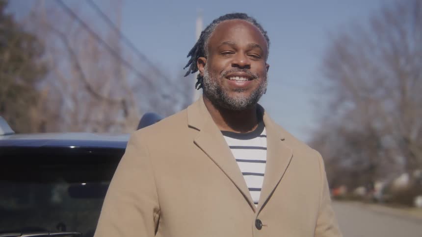 Man with dreadlocks beard and brown overcoat standing by car looks at camera smiling in the street