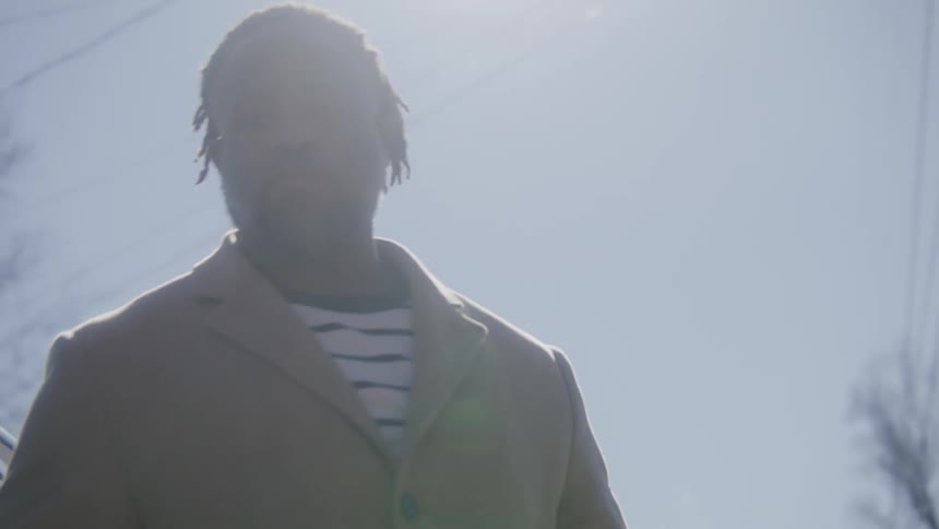 Man with dreadlocks beard and brown overcoat gets out of car in the street