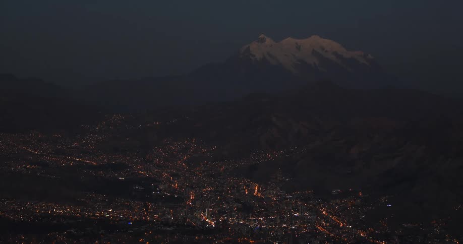 Illuminated city sits near the snow covered mountains at night