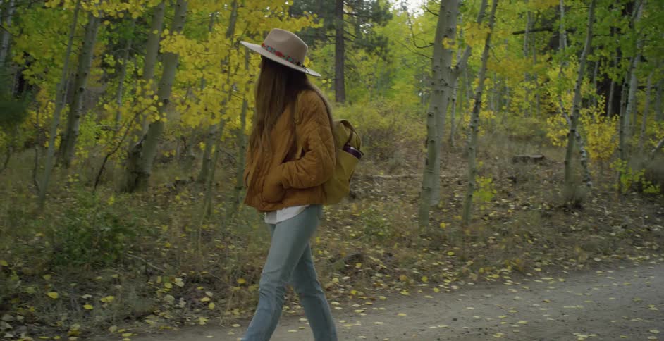 A long haired teenage girl wearing a sun hat and a backpack walks on a trail through a forest