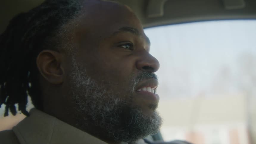 Man with dreadlocks beard and brown overcoat drives car in the street