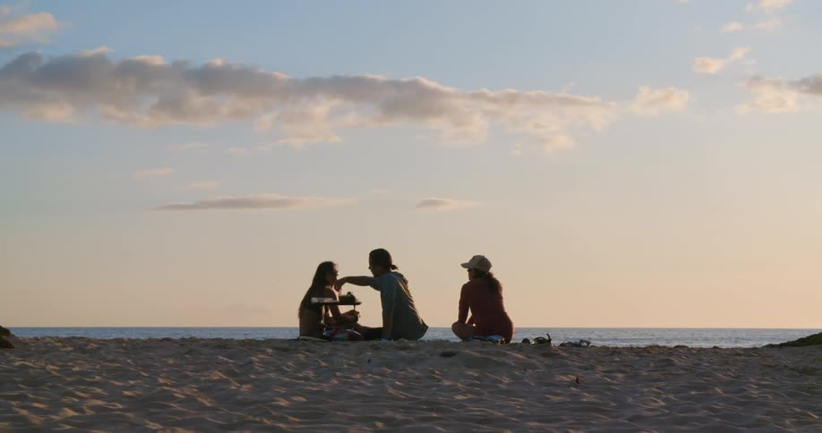 Three friends talking while sitting on the sand at the beach in front of the ocean during sunset