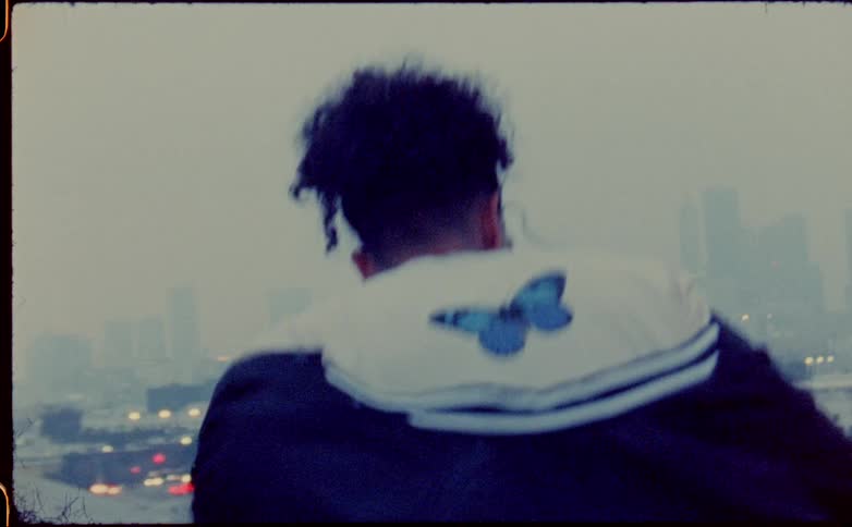 Person with black curly hair wears a heavy coat with a butterfly emblem on the hood