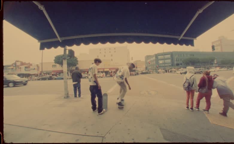 Friend watches another try a skateboard trick as traffic flows down street next to them